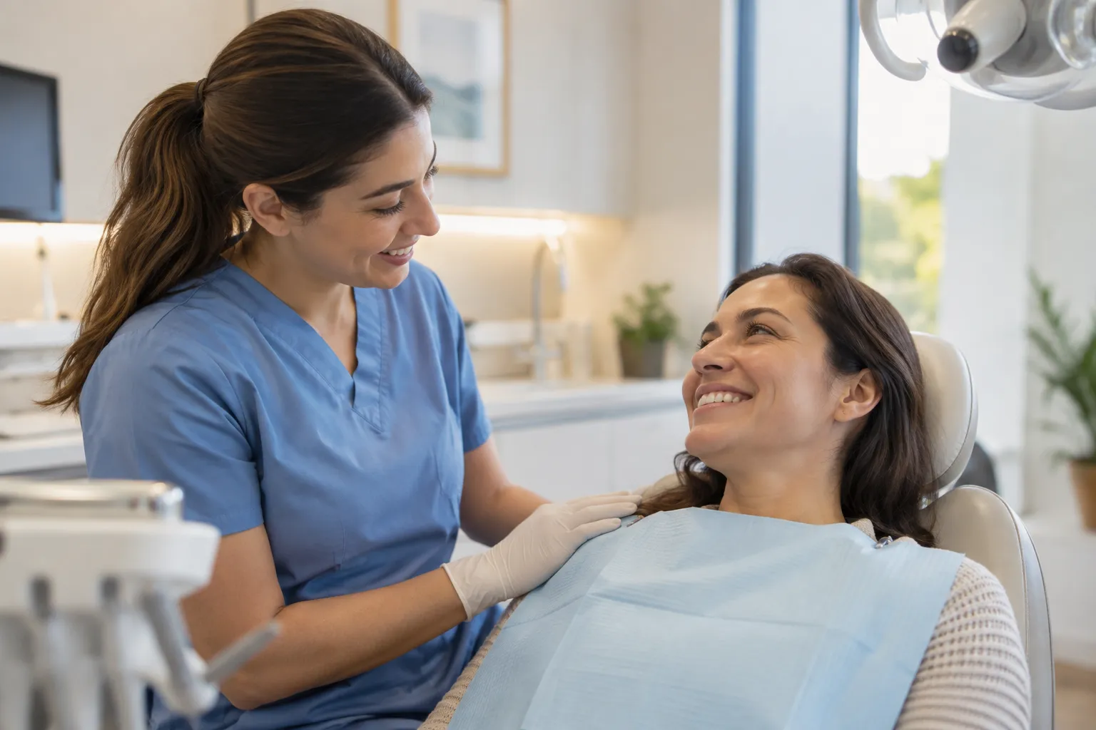 A smiling dental hygienist in blue scrubs resting a reassuring hand on a relaxed patient in a treatment chair
